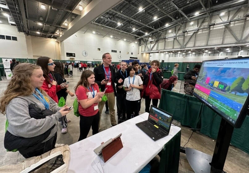 A group of people gathered around a table with a laptop and a large screen displaying a colorful quiz question in an indoor event space.